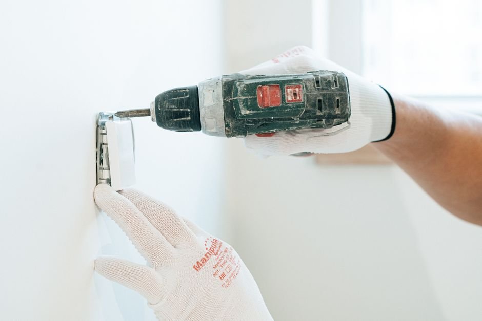 Gloved hands using a drill to install a power outlet on a wall, the kind of job a Dumbarton electrician handles